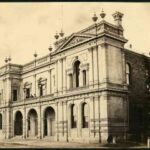 Image: A two storey building with arched windows and an arched porch supporting a balcony. Its parapet is decorated with stone urns and carved pediments.