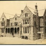 Image: A large, three-storey mid-nineteenth century bluestone building sitting atop reclaimed land. A sign reading ‘Mercantile Marine Office’ is visible at the bottom right of the building. A group of men sit on a bench near the building entrance