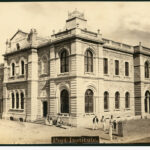 Image: An imposing, two-storey stone building in Victorian-Italianate style stands at the intersection of two dirt streets. A small group of men and boys stand in front of the building