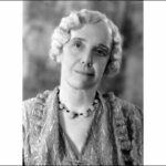 Image: A photographic head-and-shoulders portrait of a woman with medium-length wavy hari wearing 1930s attire