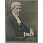 Image: A man in a barrister wig and gown poses for a photograph. He is seated in a chair with his right profile facing the camera