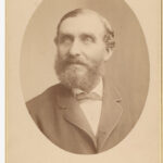 Image: A photographic head-and-shoulders portrait of a bearded middle-aged man in a suit with bowtie