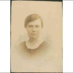 Image: A photographic head-and-shoulders portrait of a woman with ear-length dark hair and wearing 1920s attire