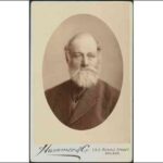 Image: A photographic head-and-shoulders portrait of an elderly, balding man with a large, bushy beard and late-Victorian era suit