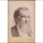 Image: A photographic head-and-shoulders portrait of a middle-aged bearded man in a suit. He has a full head of hair and is looking to the left