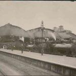 Image: a steam train is stopped beside a platform on which stands a number of men and women in early 20th century clothing. Its carriages stretch away into the distance and pass under a curved roof platform cover.