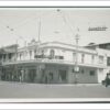 Image: a two storey corner grocery store with a self supporting verandah. The building is painted white. There are a number of pedestrians and 1950s era cars on the street outside.