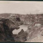 Image: A massive, steep-sided pit filled with water is flanked at the top by a number of buildings and chimneys