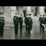 Image: A middle-aged man attired in a ceremonial dress uniform stands at the head of a group of military officers, all of whom are similarly attired. A large building with columns is visible in the immediate background