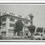 Image: a four storey white stucco building with large arched on its third floor. It sits on the corner of a tree lined street which is busy with 1950s era automobiles.