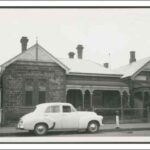 Image: a white 1950s era car is parked outside a small bluestone cottage with a verandah