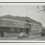 Image: a 1950s era car is parked outside of a single storey art-deco building with curved corners and glass brick windows.