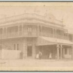 Image: a two storey corner hotel with a narrow wrap around balcony and balustraded parapet with urn decorations. On one side of the ground floor a wide verandah protrudes beyond the balcony above.  The other side is fenced off.