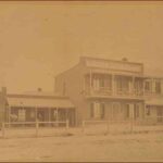 Image: a row of three terraced buildings. The one on the left is one storey with a verandah under which people stand. In the centre is a two storey building with a balcony and a parapet sign. To the right is another two storey building with a verandah.