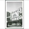 Image: 1950s era cars are parked outside a large theatre building which features a huge decorative arch inside which is a sign with the theatre's name and information about the current show. The building also features decorative plasterwork.