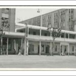 Image: a simple, two storey building with a balcony/verandah combination running the entire length of the façade is sandwiched between two much larger, more ornate buildings.