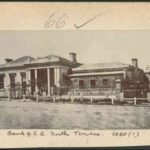 Image: a single storey building with grand columned portico under which figures can be seen standing. A garden can be seen behind a fence to the right of the building.