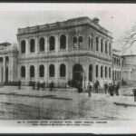 Image: a two storey square stone building on a street corner. Men in dark suits are gathered outside. The doors and windows of the building are arched and it has a flat roof with a balustraded parapet.