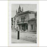 Image: a girl stands outside a simple two storey stone building with protruding wooden balcony and a sign reading "Queen's Chambers". Behind the building the spires of a small church can be seen.