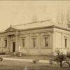 Image: Old photograph of a single storey building with a door and six window along the front and a single window visible on the adjacent side of the building.  The building also features a small portico and is reasonably ornate.