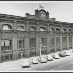 Image: 1960s cars are parked outside a large, three storey brick factory building. Some of the windows bear striped awnings and there is a parapet sign reading "Fowlers Lion Factory" which is topped with a life-sized lion statue