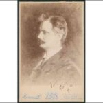 Image: A photographic head-and-shoulders portrait of a young man wearing a late-Victorian era suit and sporting a dark, handlebar moustache