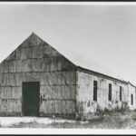 Image: A simple, single-storey building clad in metal sheeting stands in an open scrubby area