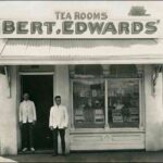 Image: Two men stand in the doorway of a small shop building. A sign on the roof of the shop reads ‘Tea Rooms, Bert.Edwards’