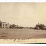 Image: a wide dirt road with a scattering of one to three storey stone and brick buildings. Parked outside one of the buildings is a line of horse drawn vehicles for hire.