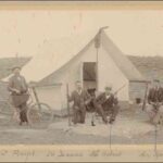 Image: Four men sit on chairs in front of a tent. One man holds a bicycle while two others brandish rifles