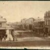 Image: a view of a city street which is lined with two and three storey commercial buildings. A large number of male and female pedestrians in 1860s attire can be seen walking down the sidewalks or crossing the road.