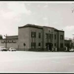 Image: A two-storey building with plain façade at the intersection of two streets. 1960s-era auto-mobiles are driving on the street in front of, and adjacent to, the building