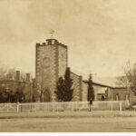 Image: A low, squat church building stands at the intersection of two dirt roads. Two people stand in front of the church