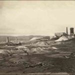 Image: Several large chimneys and buildings are arrayed among numerous tailings piles. A low mountain range is visible in the background