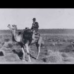 Image: A man wearing a fedora and suit jacket rides a camel in a flat expanse of scrubby desert