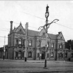 Image: A two-storey brick building with ornate facade is located at the corner of two dirt roads. A lamppost and tram tracks are visible in the foreground