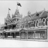 Image: a two storey ornate brick building with arched windows, a series of store windows beneath a verandah, two towers with square cupolas and flagpoles flying Australian flags above the main entrance and a tiled roof