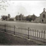 Image: A large stone church and stone house stand adjacent to one another next to a dirt street. Part of an iron fence is visible in the immediate foreground