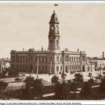 Image: a large two storey stone public building with a tall clock tower protruding from one corner stands on the corner of two city streets. A large park can be seen in the foreground of the image.