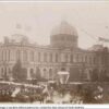 Image: a large crowd of people in 1880s clothing gather in front of a large building with a domed roof watching as horse drawn carriages pass by.