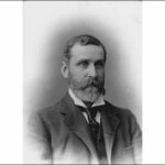 Image: A photographic head-and-shoulders portrait of a bearded, middle-aged man wearing a suit and tie