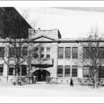 Image: a symmetrical fronted, two storey stone building with large rectangular windows and a central arched entranceway. Above the front door is a small balconet