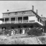 Image: A large, three-storey stone stately home with wraparound second-storey verandah. A group of people are gathered outside at the front of the house