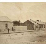 Image: men and boys in 1860s attire stand on a paved sidewalk outside their cottages.