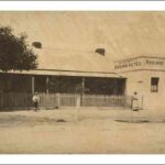 Image: a single storey cottage with pitched roof and verandah situated behind a picket fence adjoins a small square, flat roofed building with a central door flanked by two small windows. The square building has signs reading: "Railway Hotel"