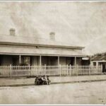 Image: A group of boys are sitting on the kerb in front of terraced cottages with a white picket fence. Geese can be seen near a tumbledown building on the right