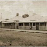 Image: a row of three identical brick, iron-roofed cottages with fenced in verandahs. A group of four children stand with their mother outside the rightmost cottage. On the far left is a fourth building which is being used as a general store.