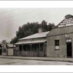 Image: a corrugated iron workshop with a gable roof and a central doorway flanked by a single window on either side. Next door is a small cottage with a verandah and central chimney.