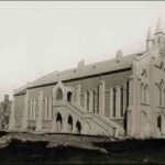 Image: a side view of a stone church with simple facade including three short spires placed centrally at one end of the building. A large staircase runs up the side of the church providing access to a second storey entrance.