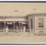 Image: A small group of men and women stand in front of a one-storey stone house with a covered verandah. A white picket fence separates the house from a dirt street